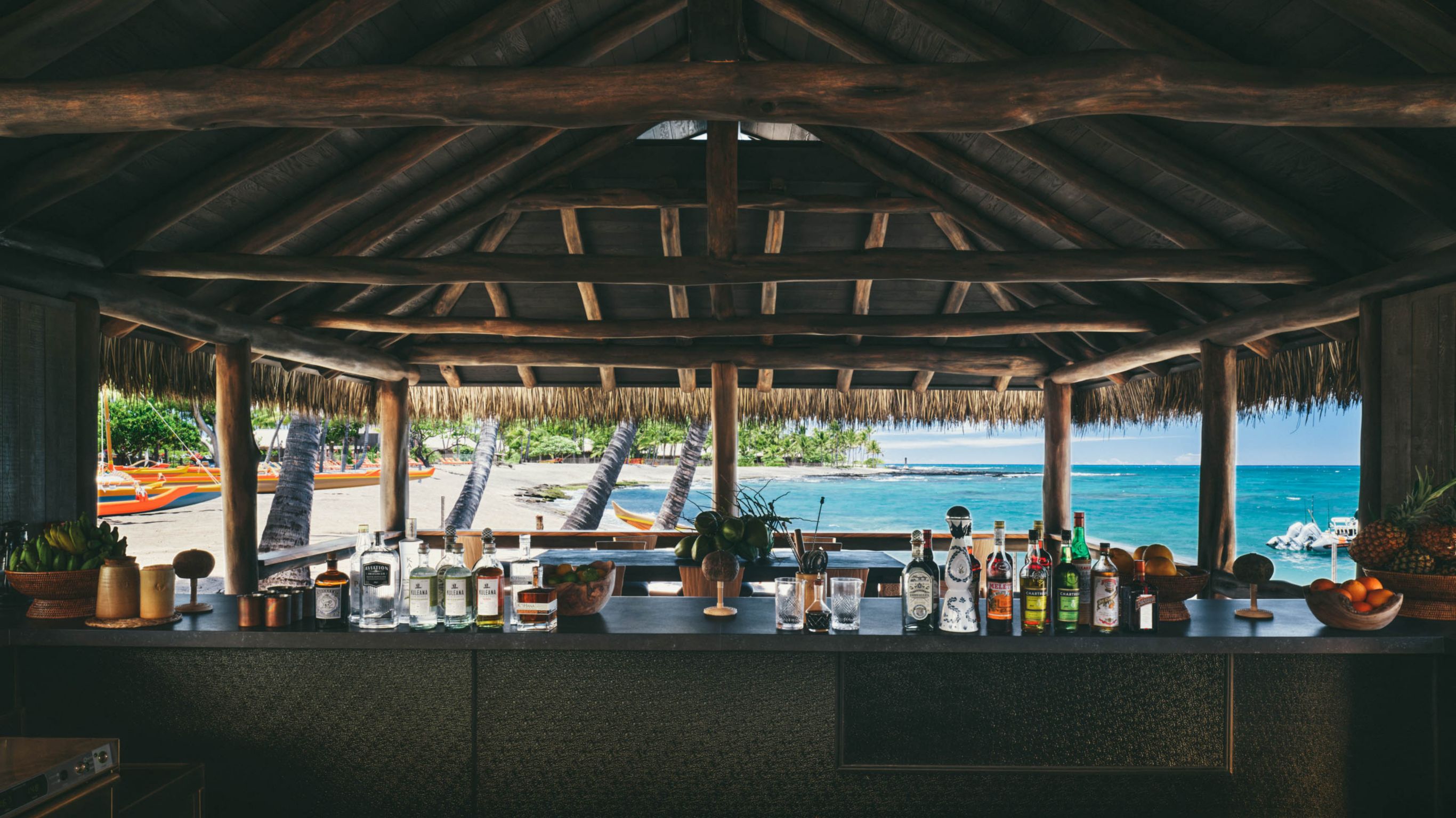 A beach bar in Hawaii with a thatched roof featuring various bottles and fruits on the counter, overlooking the ocean.